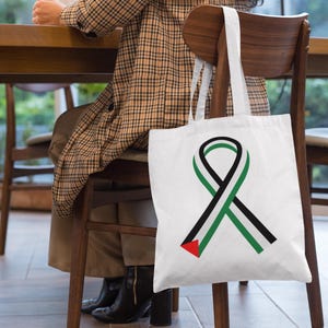 Op de afbeelding: Witte draagtas met een zwart en groen lintontwerp en een rode driehoek. De tas hangt aan een houten stoel. De persoon op de foto draagt een geruite jas.