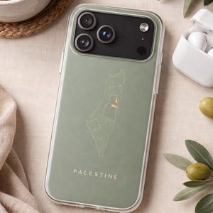 以下が含まれることがあります： パレスチナの地図と国名が白で描かれた、透明な縁取りのあるセージグリーンの携帯電話ケース。携帯電話ケースは、コーヒーカップ、オリーブ、ワイヤレスイヤホンのペアの隣にあります。