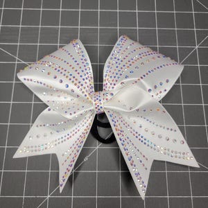 May include: White satin cheerleading bow adorned with iridescent rhinestones. The bow features a classic bow shape with multiple layers and is attached to a black elastic band. The rhinestones are arranged in lines across the bow.
