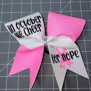 May include: A cheerleading bow with white and pink glittery ribbons. The bow features the text "In October We Cheer" and "for Hope" with a pink ribbon graphic, likely for breast cancer awareness.