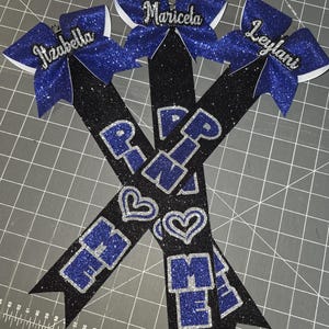 May include: Three cheerleading bows with glitter accents. Each bow has a name: Izabella, Maricela, and Leylani. The black ribbons have the letters "R", "M", and "E" in blue glitter, with silver glitter hearts.