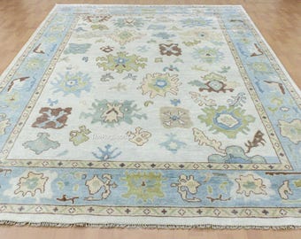 May include: Rectangular area rug with a light blue border and a cream-colored center. The rug features a repeating pattern of floral and geometric shapes in shades of blue, green, and brown. The rug is placed on a wooden floor.