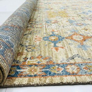 May include: Rolled-up area rug with a beige, blue, and orange pattern. The rug features a detailed border and intricate central design. The rolled section reveals a blue and brown patterned edge. The text "TheRugLoom" is visible on the rug.