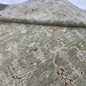 May include: A sage green area rug with an intricate, repeating pattern in shades of brown, gray, and cream. The rug is folded over, revealing the detailed border design. The texture appears soft and plush, suitable for a living room or bedroom.