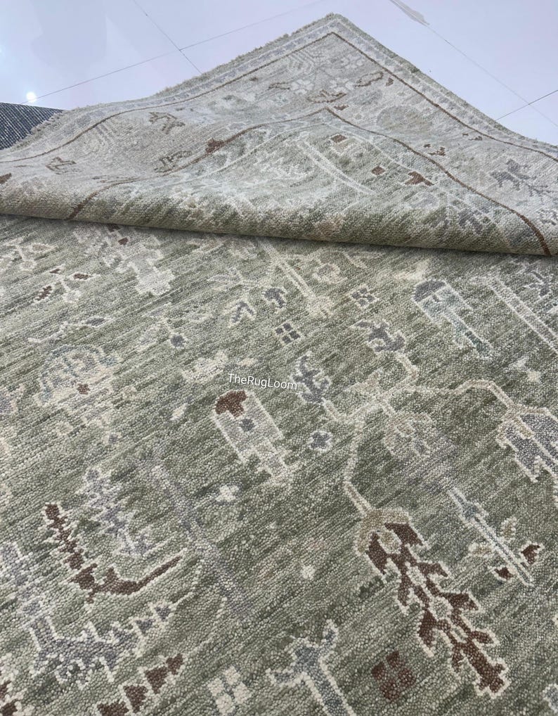 May include: A sage green area rug with an intricate, repeating pattern in shades of brown, gray, and cream. The rug is folded over, revealing the detailed border design. The texture appears soft and plush, suitable for a living room or bedroom.