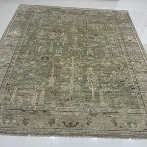 May include: A large, rectangular area rug in shades of green and beige. The rug features an intricate, repeating pattern of stylized trees and geometric shapes. The border has a darker, more detailed design with fringe along the edges.