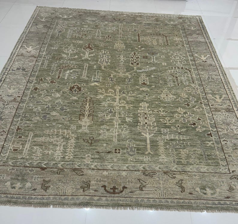 May include: A large, rectangular area rug in shades of green and beige. The rug features an intricate, repeating pattern of stylized trees and geometric shapes. The border has a darker, more detailed design with fringe along the edges.