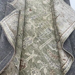 May include: A rolled-up, distressed-look rug with a faded green and gray central design. The rug features a detailed, repeating pattern with brown and cream accents. The border is a lighter color with a detailed design. The rug's edge is a dark gray.