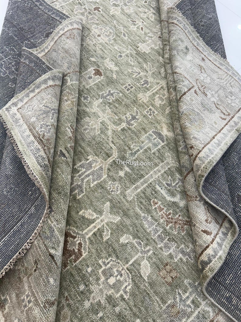 May include: A rolled-up, distressed-look rug with a faded green and gray central design. The rug features a detailed, repeating pattern with brown and cream accents. The border is a lighter color with a detailed design. The rug's edge is a dark gray.