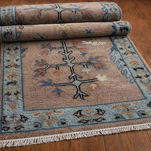 May include: A rolled-up, rectangular rug with a central tree-like design in black, blue, and orange on a tan background. The rug has a blue border with floral patterns and a fringed edge. The text "TheRugLoom" is visible.
