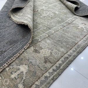 May include: A rolled-up, distressed-look rug with a muted color palette of gray, beige, and olive green. The rug features a detailed, faded pattern and a fringed edge. The text "TheRugLoom" is visible on the rug.