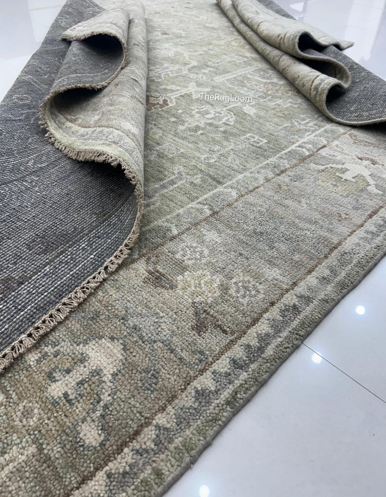 May include: A rolled-up, distressed-look rug with a muted color palette of gray, beige, and olive green. The rug features a detailed, faded pattern and a fringed edge. The text "TheRugLoom" is visible on the rug.