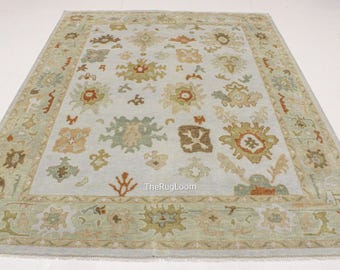 May include: A rectangular area rug with a light blue central field and a decorative border. The rug features floral and geometric patterns in shades of green, brown, and orange. The rug is made of a soft material.