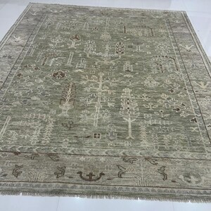 May include: A large, rectangular, sage green rug with an intricate, repeating pattern of stylized trees, floral motifs, and geometric shapes. The rug has a border with decorative elements and a fringed edge. The text "TheRugLoom" is visible.