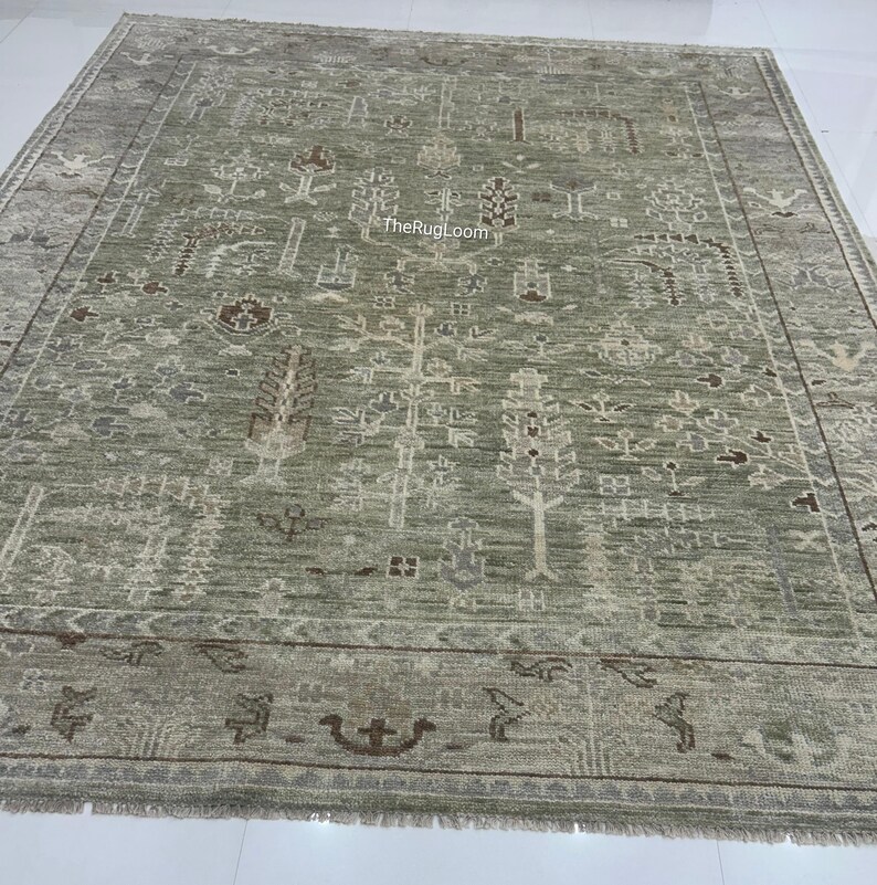 May include: A large, rectangular, sage green rug with an intricate, repeating pattern of stylized trees, floral motifs, and geometric shapes. The rug has a border with decorative elements and a fringed edge. The text "TheRugLoom" is visible.