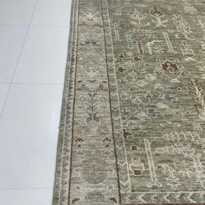 May include: Close-up of a section of a sage green and brown area rug. The rug features an intricate, faded, traditional pattern with a border. The rug is on a white tile floor. The Rug Loom is printed on the rug.