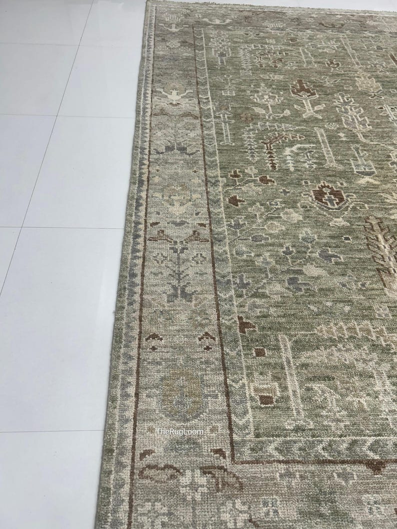 May include: Close-up of a section of a sage green and brown area rug. The rug features an intricate, faded, traditional pattern with a border. The rug is on a white tile floor. The Rug Loom is printed on the rug.