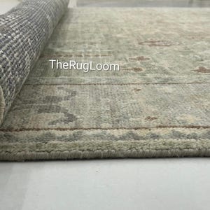 May include: A partially rolled-up area rug with a muted green and beige color palette. The rug features a traditional pattern with floral and geometric elements. The underside of the rolled portion reveals a woven backing. The text "TheRugLoom" is visible on the rug.