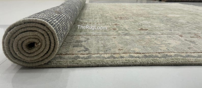 May include: A partially rolled-up area rug with a muted green and beige color palette. The rug features a traditional pattern with floral and geometric elements. The underside of the rolled portion reveals a woven backing. The text "TheRugLoom" is visible on the rug.