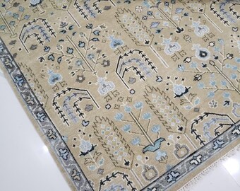 May include: Beige area rug with a detailed pattern featuring stylized trees and floral motifs in shades of blue, gray, and brown. The rug has a border with a geometric design and a fringed edge. The rug is partially rolled up.
