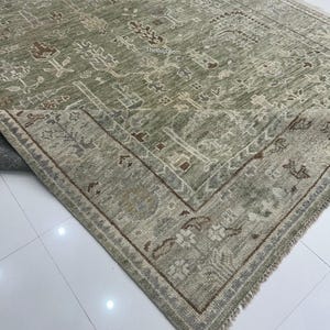 May include: A sage green area rug with an intricate, traditional pattern. The rug features a central field with floral and geometric motifs in shades of brown, beige, and gray. A detailed border frames the design. The Rug Loom is printed on the rug.