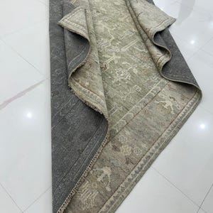 May include: A rolled-up, rectangular rug with a distressed, vintage appearance. The rug features a central field of muted green with a detailed, faded pattern. The border is a lighter shade with similar design elements. The underside is a dark gray.