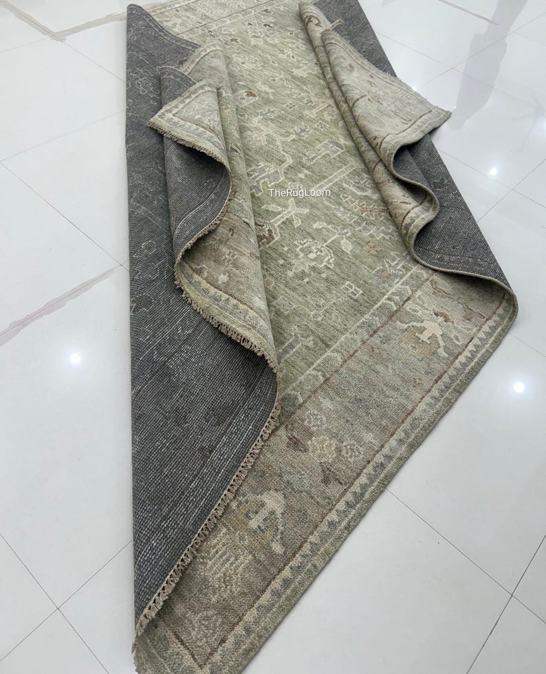 May include: A rolled-up, rectangular rug with a distressed, vintage appearance. The rug features a central field of muted green with a detailed, faded pattern. The border is a lighter shade with similar design elements. The underside is a dark gray.