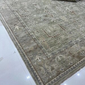 May include: A rectangular, distressed-look rug with a muted green and beige color palette. The rug features an intricate, repeating pattern with brown accents and a detailed border. The rolled-up portion reveals a dark backing.