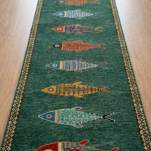 May include: A long, rectangular runner rug in a deep green hue, featuring a series of ten stylized fish motifs in various colors including red, blue, yellow, and orange. The rug has a decorative border and white fringe. Text reads TheRugLoom.
