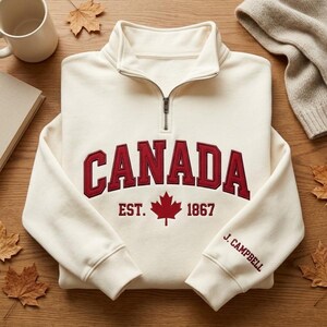 May include: A cream-colored quarter-zip sweatshirt featuring the word CANADA in large red letters, a red maple leaf, and the text EST. 1867. The left sleeve includes the embroidered name J. CAMPBELL in red, with a metal zipper at the collar.