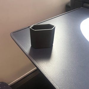 Op de afbeelding: Een zwarte, geometrisch gevormde beker met een zilveren rand staat op een donkergrijze tray. De beker heeft een achthoekige vorm. De tray maakt deel uit van een groter oppervlak, mogelijk een tafel of bureau.