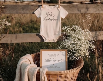 Pode incluir: Um body de bebé cor de creme com o texto Baby Anderson impresso a preto, pendurado sobre um cesto de vime contendo uma placa branca que diz We've Been Keeping A Little Secret Arriving November 202X e uma manta de malha cor de creme.