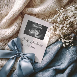 May include: A pregnancy announcement card with a sonogram image, the text "Baby Hudson" and "JUNE 2026". The card is placed on a knitted blanket with a blue satin ribbon and a bouquet of white flowers.