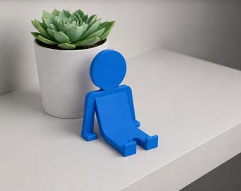 May include: A bright blue phone stand shaped like a stylised human figure in a seated position with arms extended backwards. Next to the stand is a small green succulent plant potted in a simple white cylindrical container with a smooth finish.