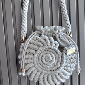May include: A cream-colored macrame bag with a circular design and a drawstring closure. The bag has thick, braided rope handles and a small gold-colored tag that reads "YULIYA SA". Wooden beads accent the drawstring.