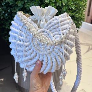 May include: A white macrame handbag with a drawstring closure and pearl accents. The bag features a round shape with a braided handle and decorative pearl strands. The bag is held in a person's hand.