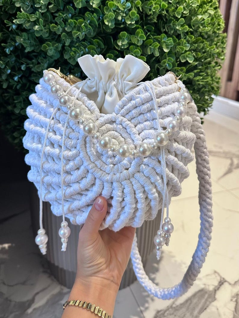 K&ouml;nnte beinhalten: Eine wei&szlig;e Makramee-Handtasche mit Kordelzugverschluss und Perlenakzenten. Die Tasche hat eine runde Form, einen geflochtenen Henkel und dekorative Perlenschn&uuml;re. Die Tasche wird in einer Hand gehalten.