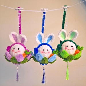 Könnte beinhalten: Drei hängende Osterhasen-Ornamente. Jeder Hase hat einen weißen Garnkörper, rosige Wangen und ein lächelndes Gesicht. Sie sind von bunten Filzblumen und Karotten umgeben. Die Hasen hängen an einer weißen Schnur mit dekorativen Stielen.