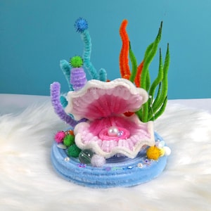 May include: A handcrafted seashell diorama with a pearl inside. The diorama features a light blue base, a pink and white shell, and colourful pipe cleaner seaweed and coral. Decorative elements include small pearls and pom-poms.