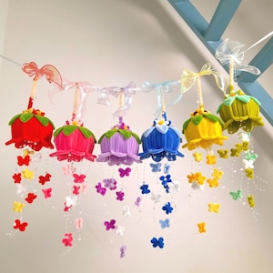 May include: A garland of six colorful, bell-shaped ornaments. Each ornament is a different color, including red, pink, purple, blue, yellow, and green. They are adorned with green leaves, white flowers, and dangling butterfly accents. Ribbons and bows are attached.