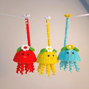 May include: Three handmade jellyfish ornaments in red, yellow, and blue, each with a smiling face and curly tentacles. They are decorated with flowers and hang from a white string, perfect for a nursery or kids room.
