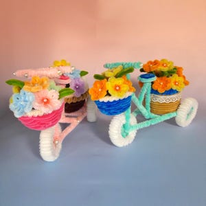 May include: Two decorative miniature bicycles made of chenille stems, each with a basket of colourful flowers. One bicycle is pink, the other is teal. The wheels are white, and the flowers are yellow, orange, blue, and pink.