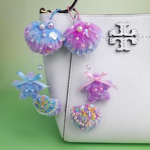 May include: A selection of handmade keyrings and bag charms in blue, purple, and pink. The charms feature seashell, star, and heart shapes, decorated with pearls, sequins, and ribbons. They are attached to a white handbag.