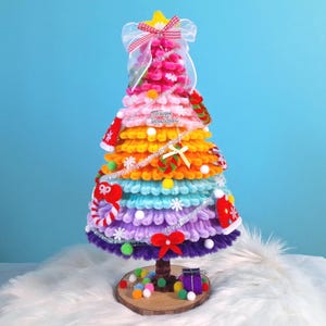 May include: A handmade Christmas tree in a rainbow of colours, with a star on top and a wooden base. The tree is decorated with ornaments, a bow, and the text "Merry Christmas". The tree is approximately 25 cm tall.