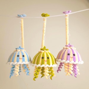 Può includere: Tre ornamenti a forma di medusa sorridente fatti a mano, blu, verdi e viola. Ognuno ha una cupola a strisce, tentacoli ricci e una parte superiore decorativa. Sono appesi a un filo bianco, perfetti per una cameretta.