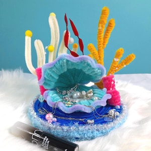 May include: A handmade seashell-shaped jewellery holder in turquoise, pink, and blue, with faux pearls and silver chains inside. Decorative coral-like elements in yellow, white, and red surround the shell. A black tube of "LASTING MELTING BALM" is in the foreground.