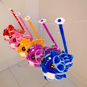 May include: A line of vibrant, plush fish decorations hanging from a white string. The fish are in various colours: red, pink, yellow, purple, and blue, each with decorative features and a flower at the top. The ornaments are suspended against a white wall.