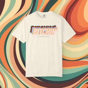 Op de afbeelding: Crèmekleurig T-shirt met het woord "BITCHIN" in gelaagde, kleurrijke tekst. Onder het woord staat het woord "THREADS" in een kleinere letter. De achtergrond heeft een retro, wervelend patroon in tinten oranje, bruin, groen en blauwgroen.