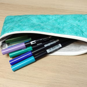 May include: A teal quilted pencil case with a white zip, partially open to reveal colourful markers. The markers include shades of green, purple, blue, and black. The case has a small tag on the side.