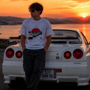 May include: A white short-sleeve t-shirt featuring a graphic print of multiple car rear ends arranged in a row under a red circle, with the text GT-R below. The shirt is worn with dark gray cargo pants and white sneakers, standing by a white sports car.